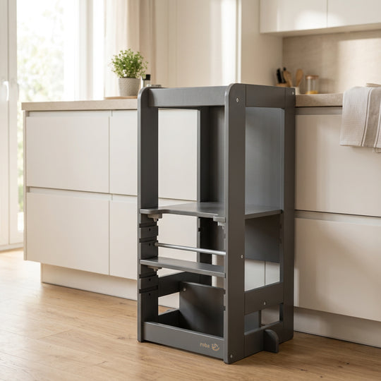 Torre de aprendizaje 'Step & Play' en Antracita, vacía en una cocina luminosa junto a una isla de cocina blanca con un paño de cocina y una planta verde sobre la encimera.