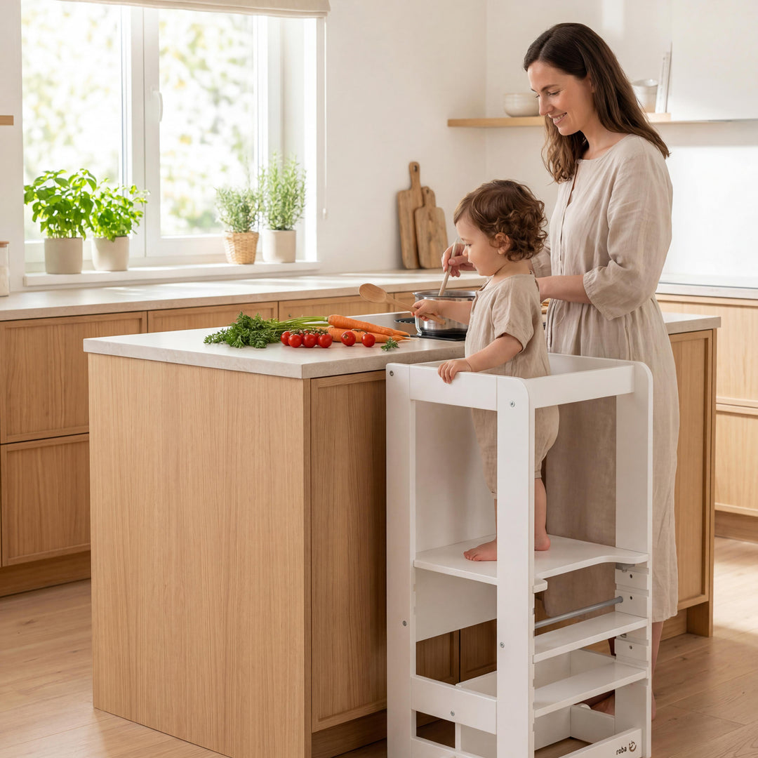 Madre e hijo pequeño de pie junto a una isla de cocina, el niño está de pie en la torre de aprendizaje blanca y ayuda a preparar verduras.